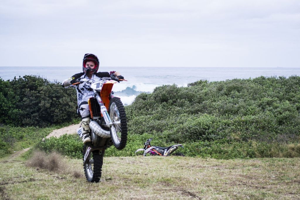 Woman dirt biker popping a wheelie on grass with the sea or ocean in the background